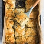 A pan of vegan spanakopita, flakey pastry pie with spinach stuffing, with a square taken out of it. The square is on a spoon, showing the layers of pastry and spinach.