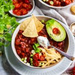 A bowl of chilli with tortilla chips, vegan cheese, vegan sour cream and an avocado on top.
