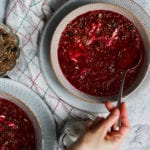 Two bowls of beetroot and tomato soup with a hand and spoon.