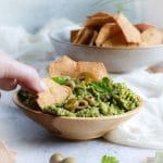Avocado and olive dip in a bowl with a hand picking up a cracker and scooping up the dip.