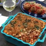Vegan Greek style 'Feta' casserole in a blue dish on a table with wine and side dish in the background