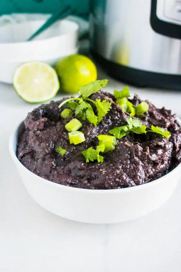 A bowl of refried black beans ready to serve. There is some cilantro on top and lime wedges in the background.
