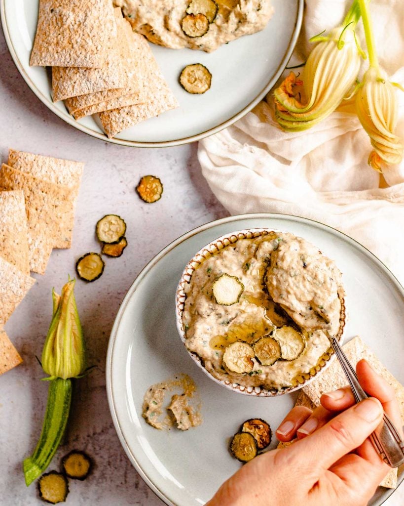 Two plates of zucchini babagaboush with crackers. A hand is reaching in to scoop up the dip.