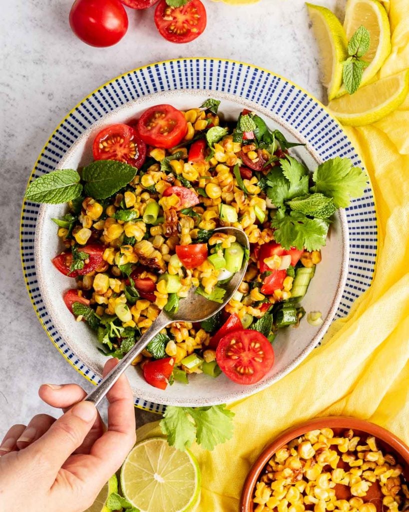 A hand reaching in with a spoon to scoop up a spoonful of the grilled corn and tomato salad.