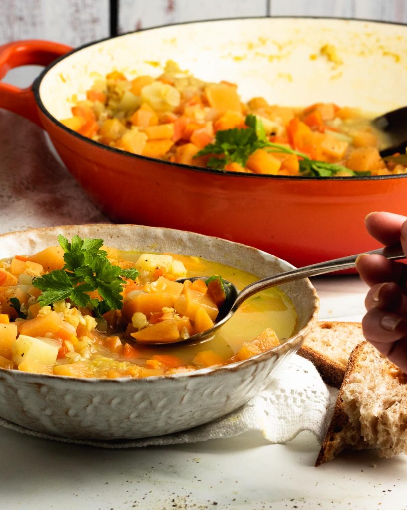 A bowl of soup with a hand coming in to scoop it up. There is a large pan of swede soup in the background too.