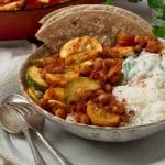 A bowl of courgette curry with rice, raita and a folded paratha bread.