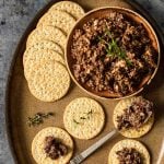 A little bowl of of mushroom tapenade on a larger plate with some crackers and a spoon.