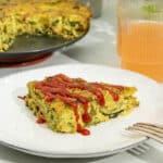 A slice of tofu frittata with vegetables that has a drizzle of sriracha sauce on top, with a glass of kombucha in the background.