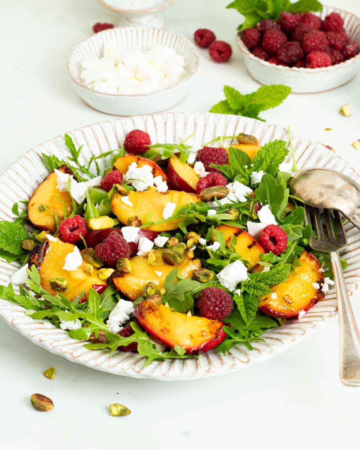 A plate of salad with grilled nectarines, arugula (rocket salad), raspberries, vegan feta and pistachio nuts.