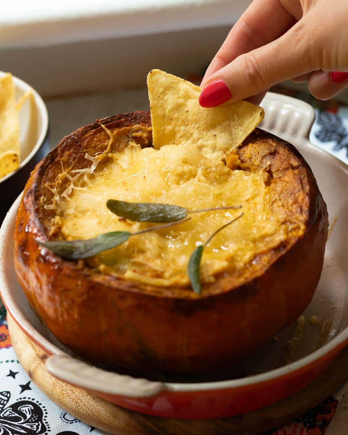 A hand holding a corn chip which is dipping into the hot cheesy dip in a whole roasted pumpkin.