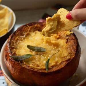 A roasted pumpkin filled with hot cheese dip with a hand reaching in with a corn chip to scoop up some dip.
