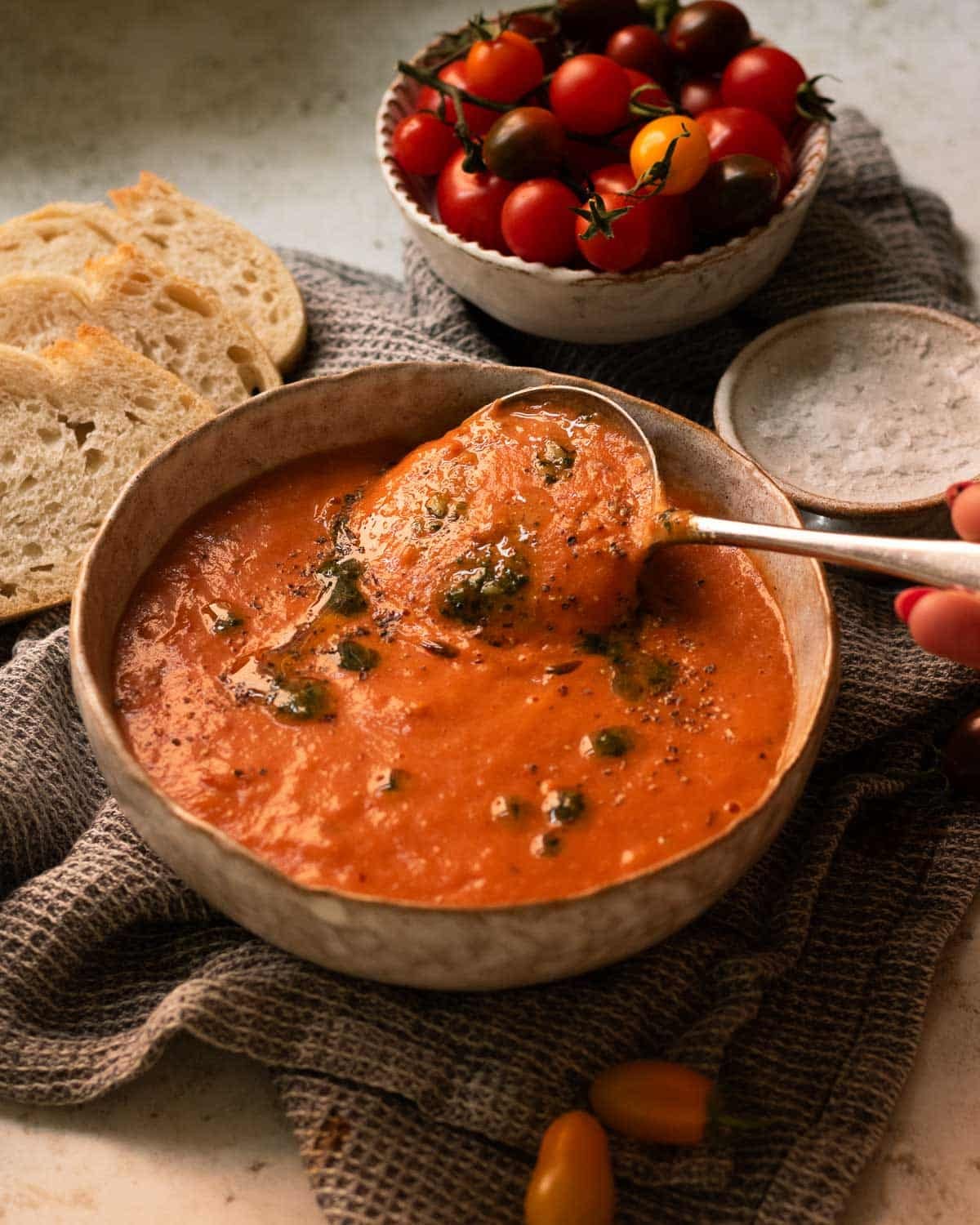 A bowl of roasted red pepper and tomato soup, ready to eat, with some bread and tomatoes in the background.