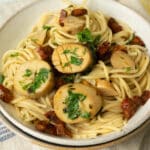 A plate of spaghetti with vegan mushroom scallops on top. Next to the plate is a glass of white wine and half a lemon.