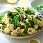 A bowl of asparagus and pea pasta salad ready to eat with some lemons in the background and a little jug of extra dressing in the foreground.