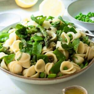 A bowl of asparagus and pea pasta salad ready to eat with some lemons in the background and a little jug of extra dressing in the foreground.