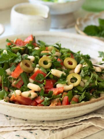 A plate of white bean salad with peppers and olives with some smaller plates and extra dressing in a pot in the background.