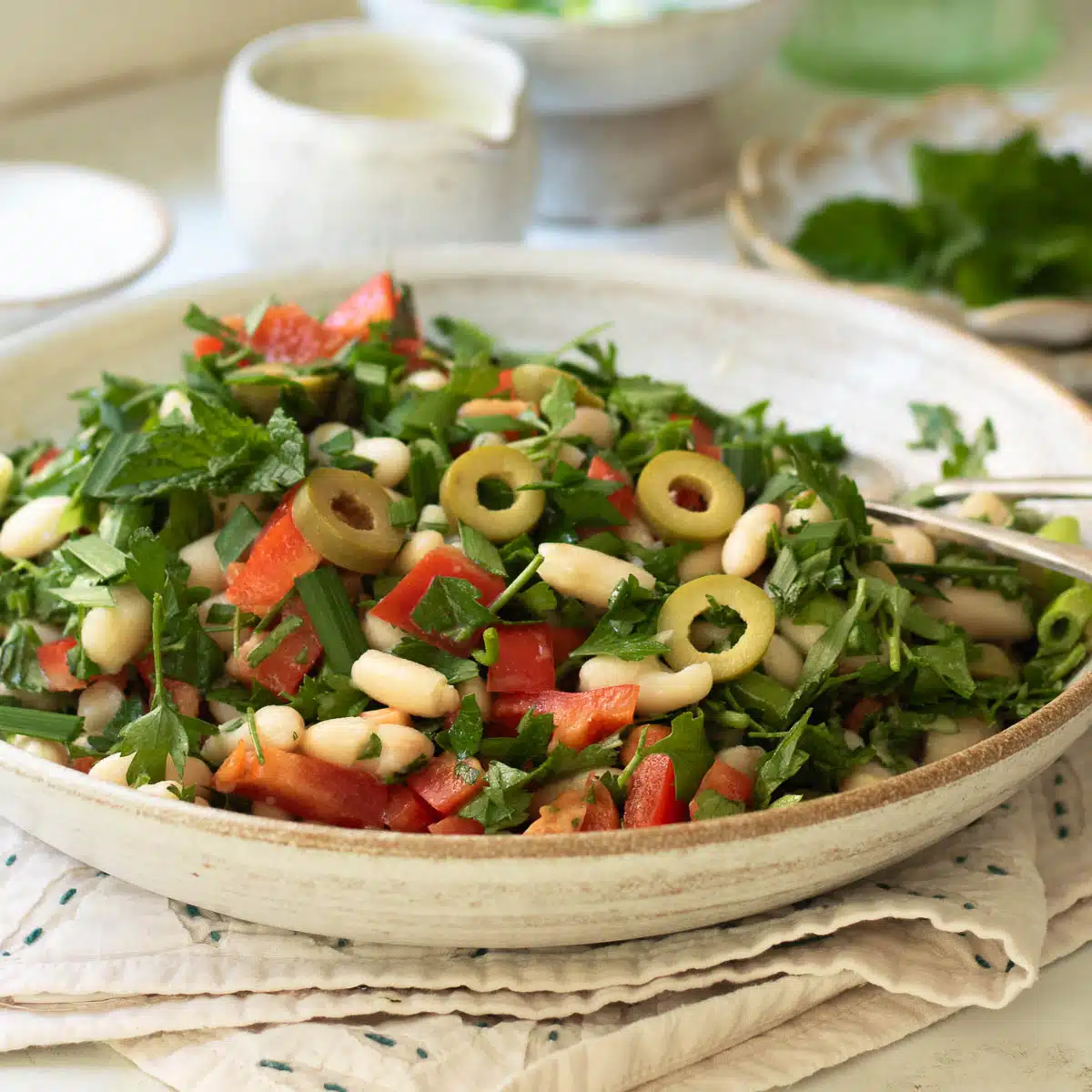 A plate of white bean salad with peppers and olives with some smaller plates and extra dressing in a pot in the background.