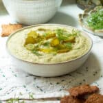 A bowl of dill pickle hummus with crackers in a bowl behind it.