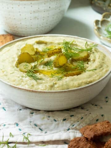A bowl of dill pickle hummus with crackers in a bowl behind it.