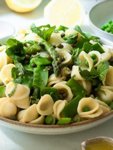 A bowl of asparagus and pea pasta salad ready to eat with some lemons in the background and a little jug of extra dressing in the foreground.