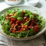 A bowl of harissa roasted chickpeas and tomatoes on a bed of arugula (rocket) with some nuts and dressing in the background.