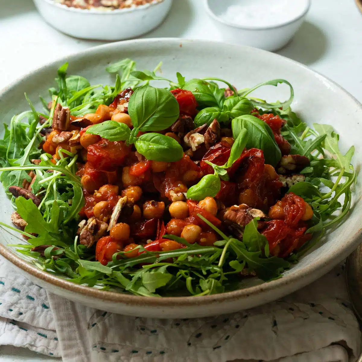 A bowl of harissa roasted chickpeas and tomatoes on a bed of arugula (rocket) with some nuts and dressing in the background.