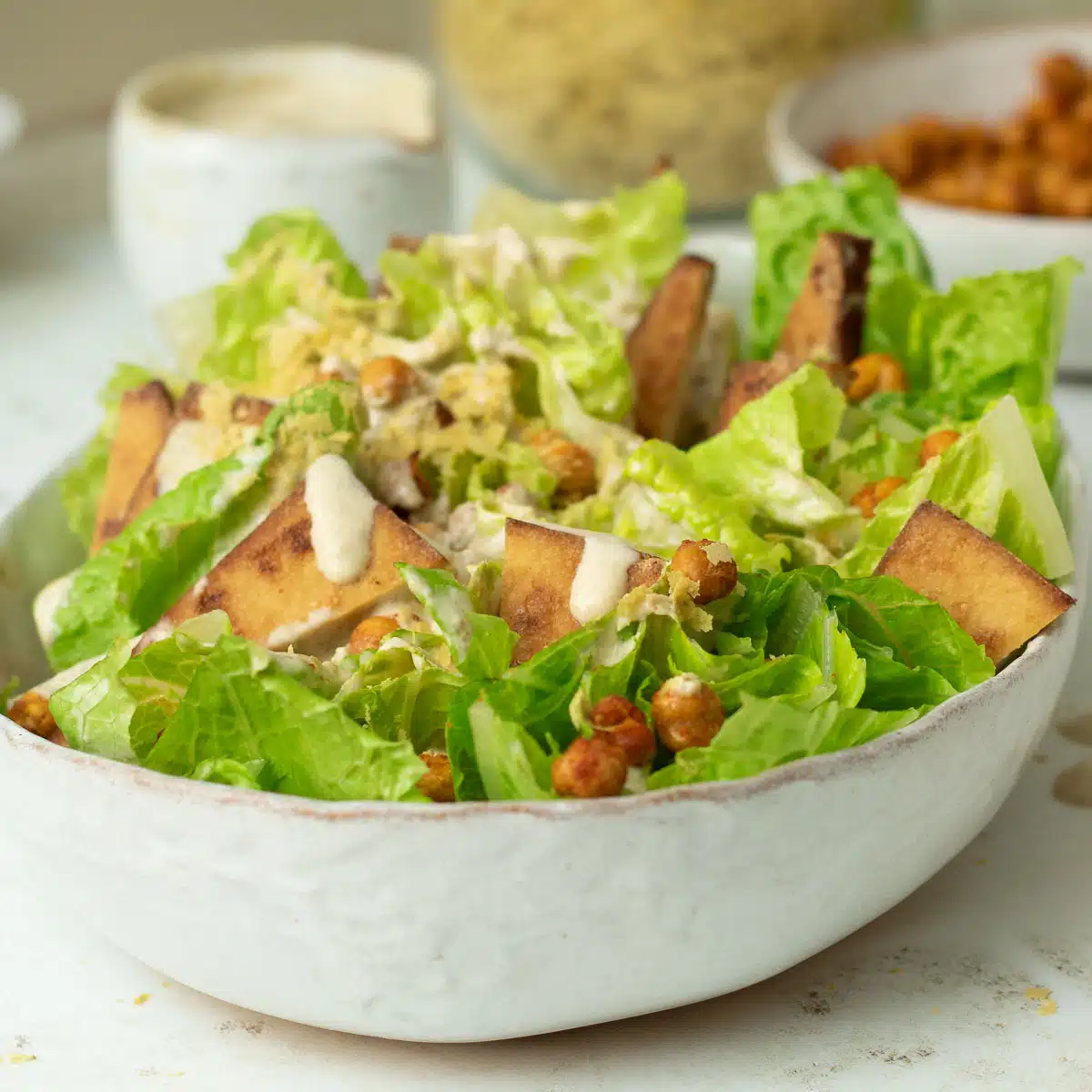 A bowl of vegan caesar salad with tahini dressing. There are crispy chickpea croutons and a jar of nutritional yeast behind the salad bowl.