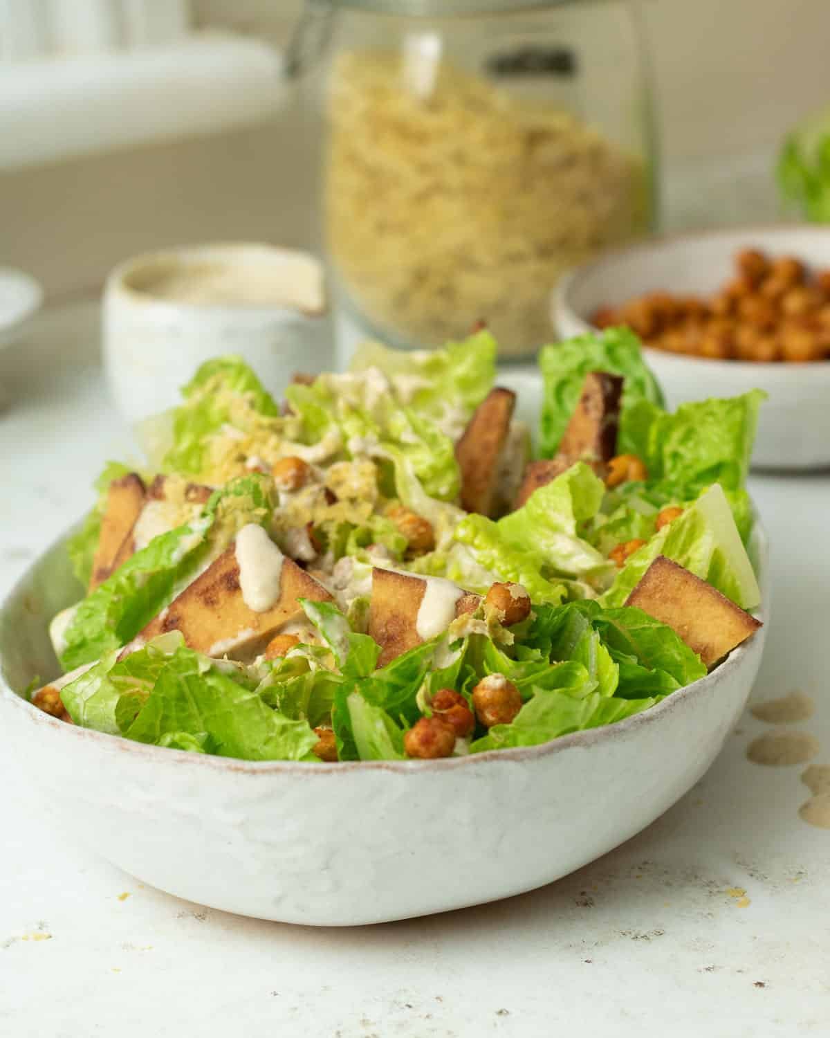 A bowl of vegan caesar salad with tahini dressing. There are crispy chickpea croutons and a jar of nutritional yeast behind the salad bowl. 
