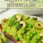 A white bowl of Green Pea Dip with Olives and extra peas on top with little leaves of mint and a drizzle of olive oil on top.