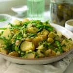 A large bowl of potato salad with peas, sugar snap peas, dill and pickles mixed through it. There is a large jar of dill pickles in the background.