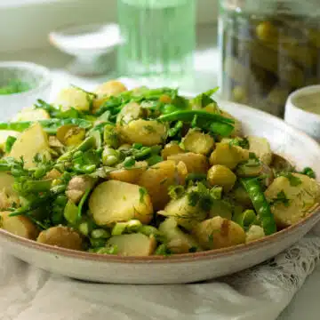 A large bowl of potato salad with peas, sugar snap peas, dill and pickles mixed through it. There is a large jar of dill pickles in the background.