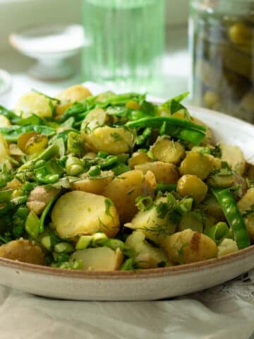A large bowl of potato salad with peas, sugar snap peas, dill and pickles mixed through it. There is a large jar of dill pickles in the background.