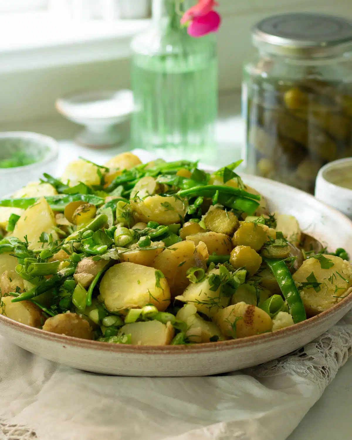 A large bowl of potato salad with peas, sugar snap peas, dill and pickles mixed through it. There is a large jar of dill pickles in the background.