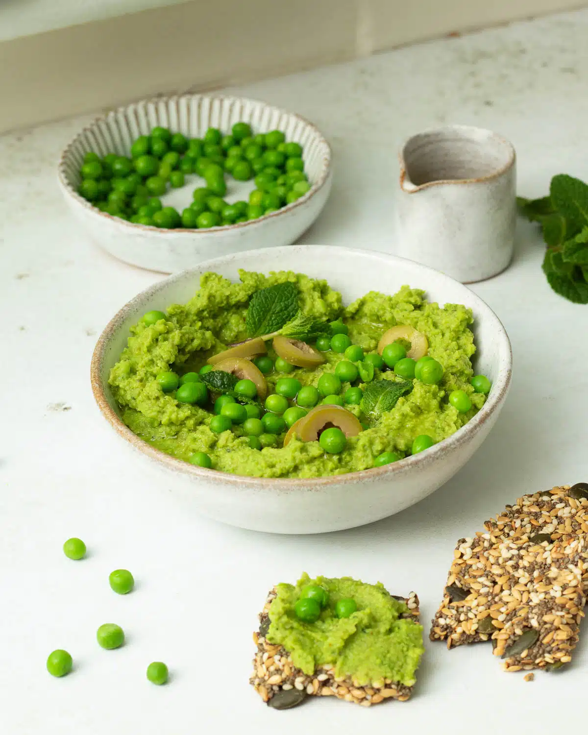 A white bowl of Green Pea Dip with Olives and extra peas on top with little leaves of mint and a drizzle of olive oil. There are some crackers in the foreground with extra dip on them. 
