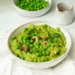 A white bowl of Green Pea Dip with Olives and extra peas on top with little leaves of mint and a drizzle of olive oil on top.