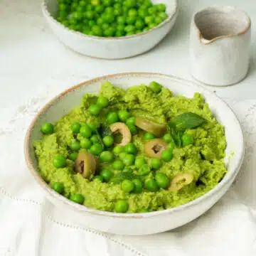 A white bowl of Green Pea Dip with Olives and extra peas on top with little leaves of mint and a drizzle of olive oil on top.