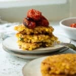 A stack of tofu and corn fritters with some roasted tomatoes on top. There is a few more fritters in the foreground.