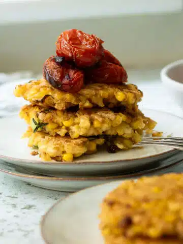 A stack of tofu and corn fritters with some roasted tomatoes on top. There is a few more fritters in the foreground.