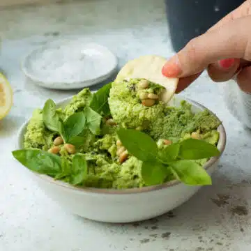 A bowl of edamame dip with basil and pine nuts on top, with a hand that has a cracker in it dipping the cracker in.