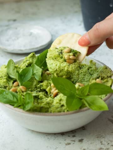 A bowl of edamame dip with basil and pine nuts on top, with a hand that has a cracker in it dipping the cracker in.