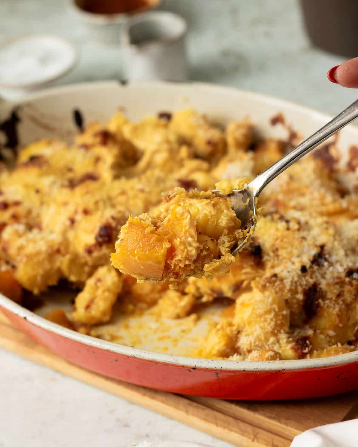 A pan of baked butternut squash and gnocchi with a cheesy harissa sauce and crunchy breadcrumb topping. There is a spoon scooping out a spoonful of squash and gnocchi.