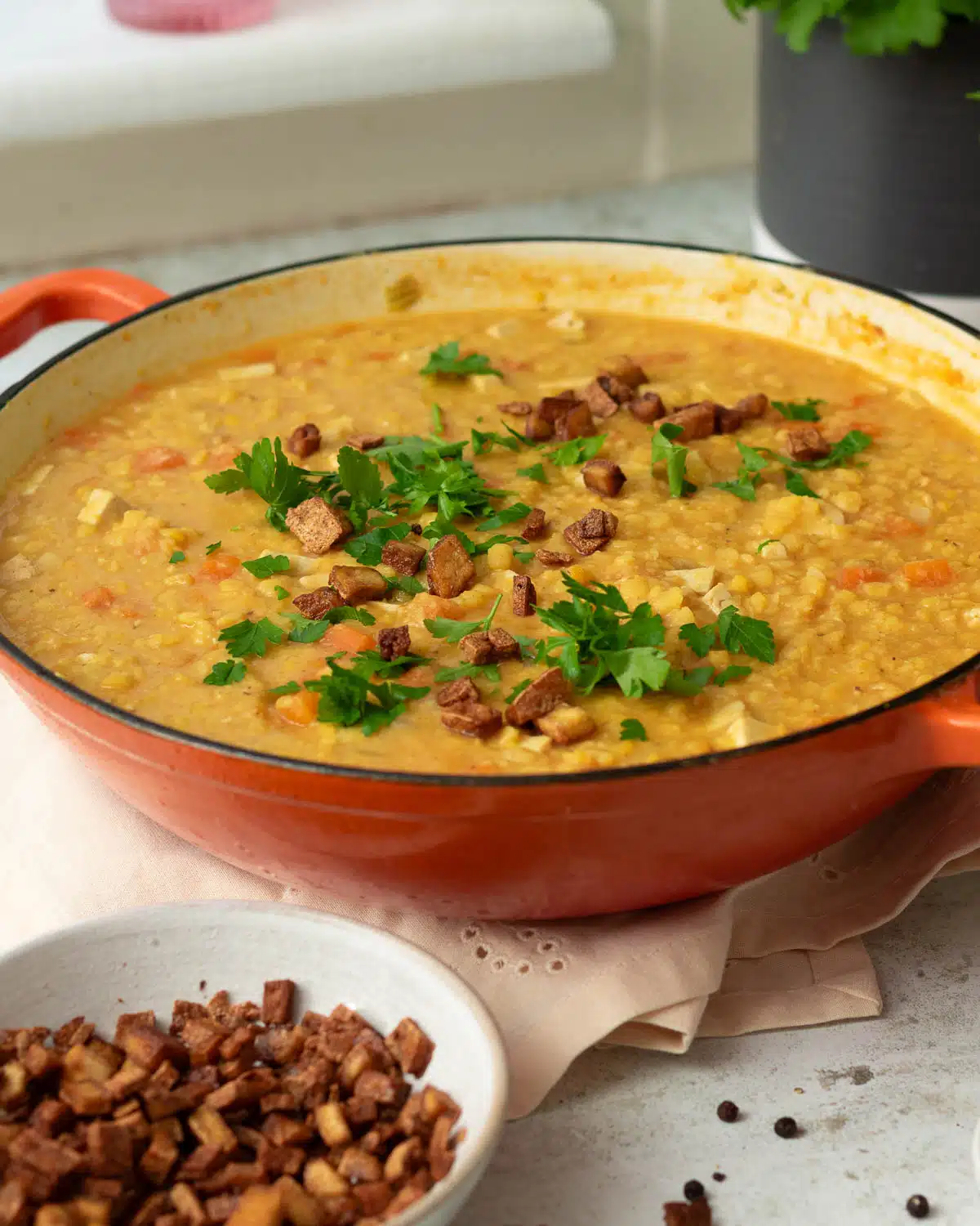 A large pan of vegan split pea and ham soup There are small cubes of vegan faux bacon sprinkled on top of the soup along with some parsley.