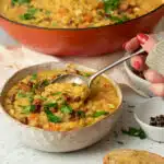 A bowl of vegan split pea and ham soup with scooping a spoonful of soup out of the bowl. . There are small cubes of vegan faux bacon sprinkled on top of the soup along with some parsley.