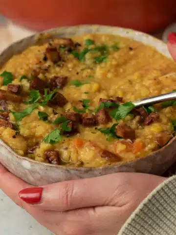 A bowl of vegan split pea and ham soup with someone holding the bowl. There are small cubes of vegan faux bacon sprinkled on top of the soup along with some parsley.