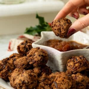 A plate of cranberry bean meatballs with dipping sauce and a hand reaching in to pick one up.