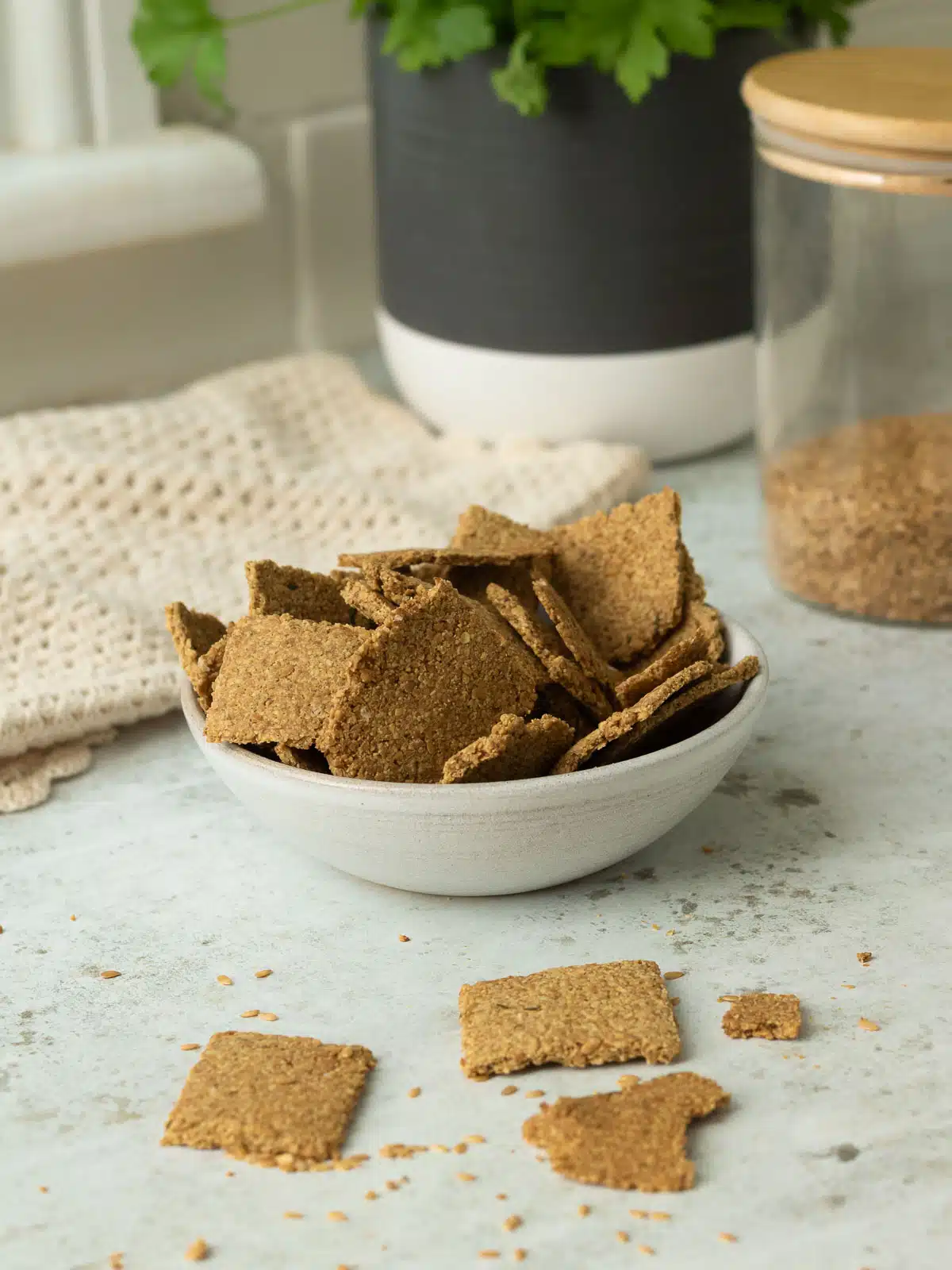 A bowl of flaxseed crackers made wthi ground flaxseed, in a bowl. There is a jar of whole flaxseeds behind it. 