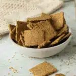 A bowl of flaxseed crackers made wthi ground flaxseed, in a bowl. There is a jar of whole flaxseeds behind it.