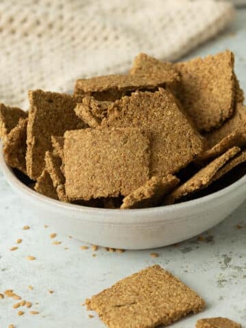 A bowl of flaxseed crackers made wthi ground flaxseed, in a bowl. There is a jar of whole flaxseeds behind it.