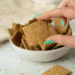 A hand picking up a single flaxseed cracker with a bowl of crackers behind.
