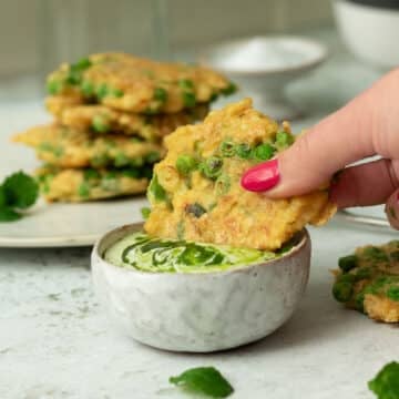 Image of a pea and tofu fritter being dunked in a creamy dipping sauce. There is a stack of fritters behind on a plate.